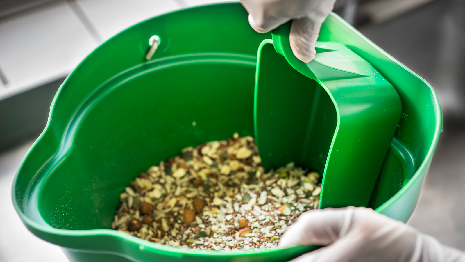 Hand met handschoen schept een mengsel van zaden of granen uit een groene emmer met een schep.