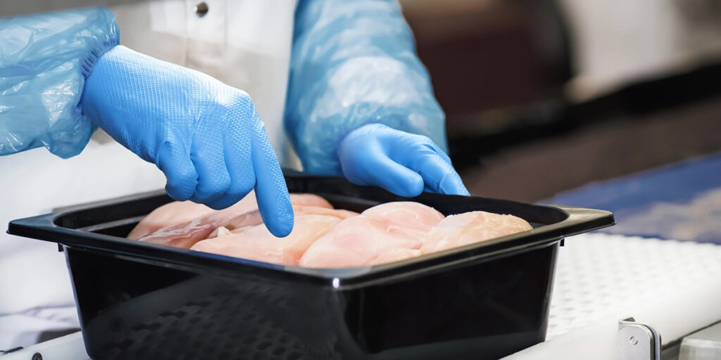 Een afbeelding van 2 handen met blauwe handschoenen boven een bak met rauwe kipfilets