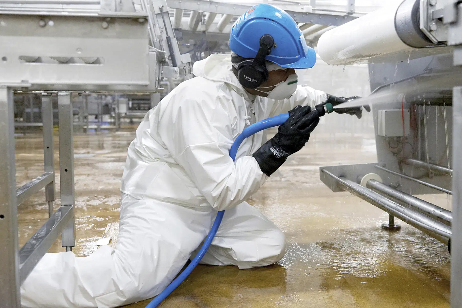 Medewerker in witte beschermende kleding reinigt industriële apparatuur met een hogedrukspuit.