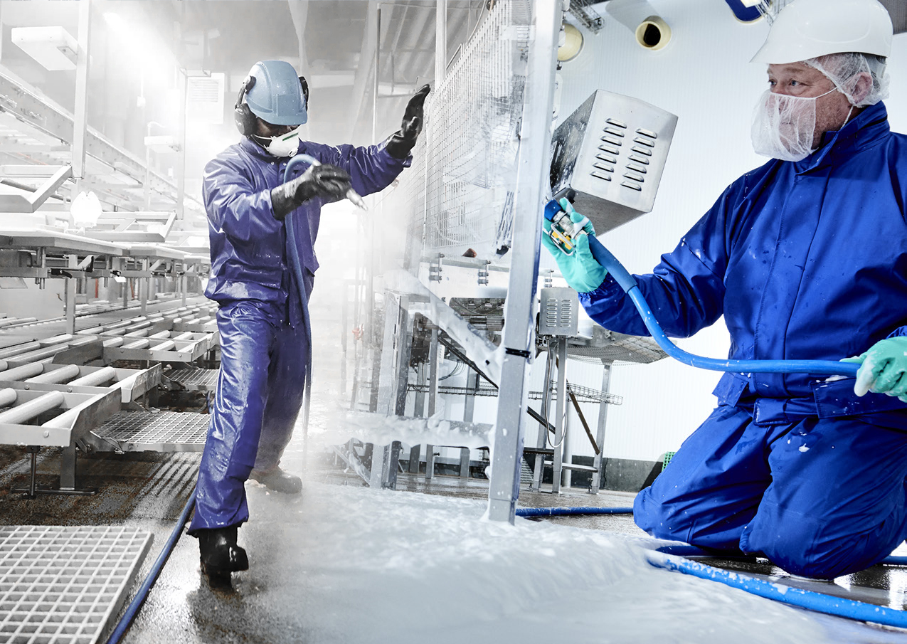 Twee medewerkers in blauwe beschermkleding reinigen machines en vloeren in een industriële productieomgeving.