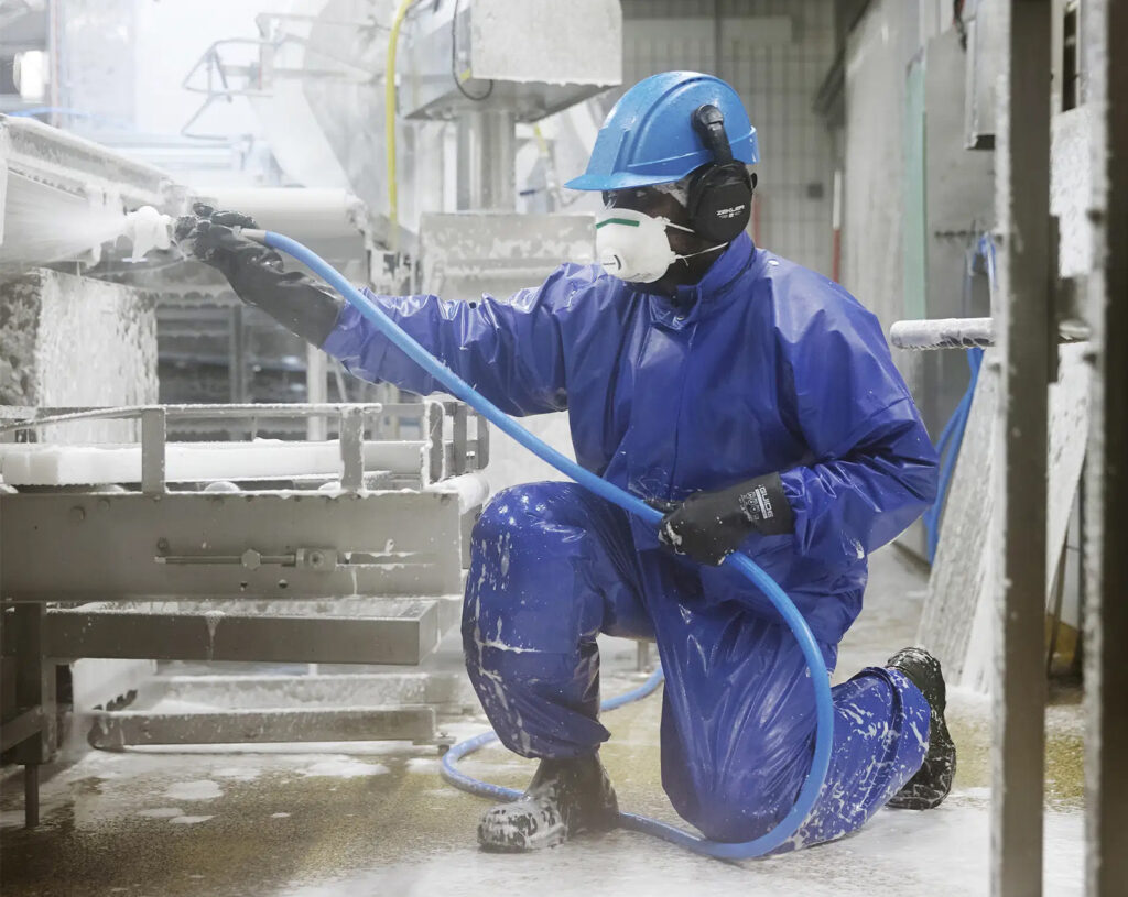 Medewerker in blauwe waterdichte beschermkleding reinigt een industriële installatie met een waterslang.