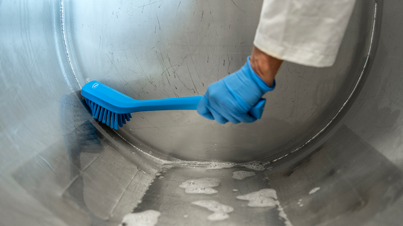 A stainless-steel round object is being cleaned with a blue Vikan brush.