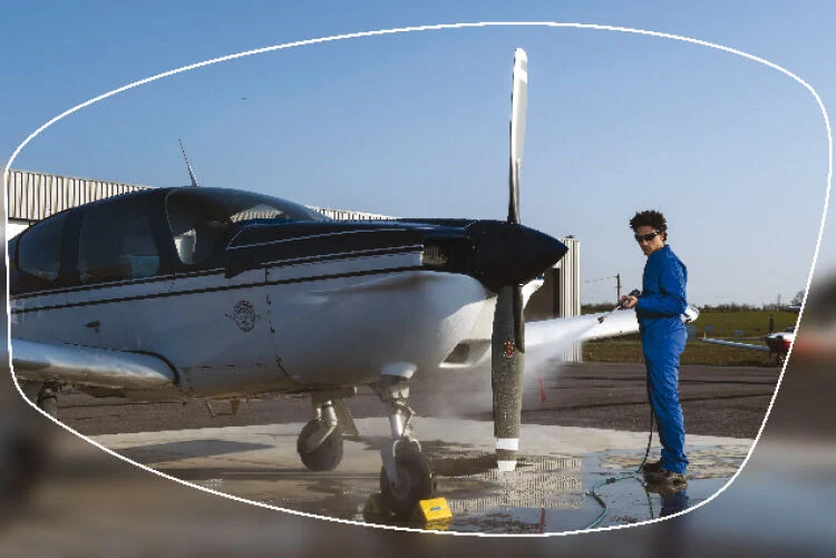 A person in blue overalls cleaning a small aircraft with a pressure washer, viewed through a protective safety lens