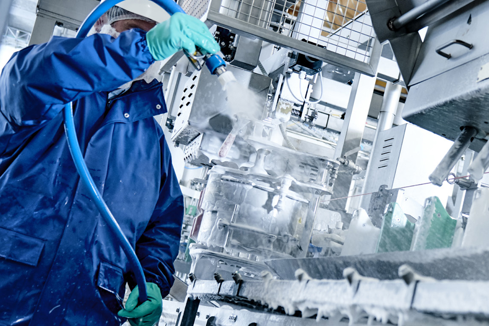 Worker wearing blue protective clothing and green gloves cleaning industrial machinery with a water hose.
